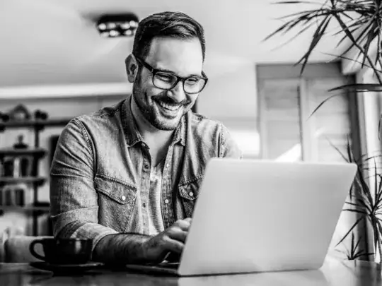 Man with glasses smiling while typing on a laptop at home.