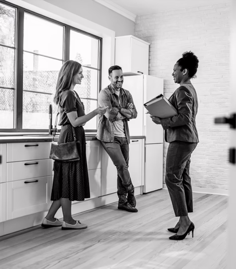 Man and woman standing in a room smiling while speaking to a real estate agent who is holding a notebook.