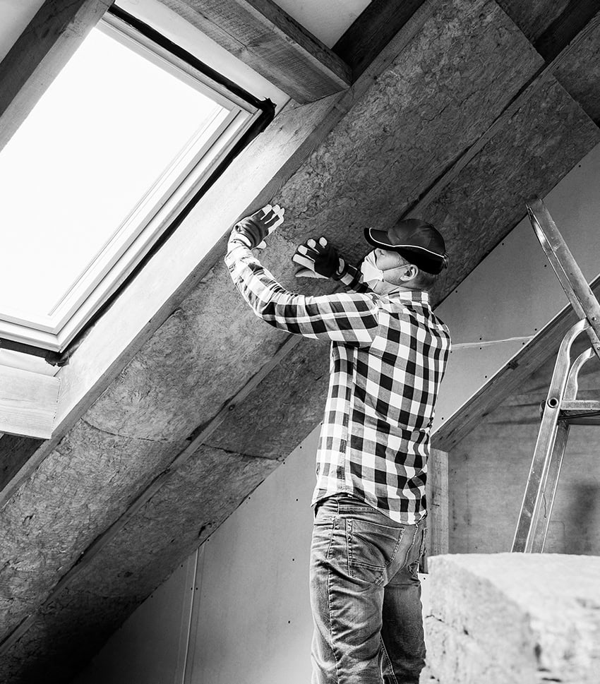 Construction worker installing insulation in attic space, using safety gloves and mask.