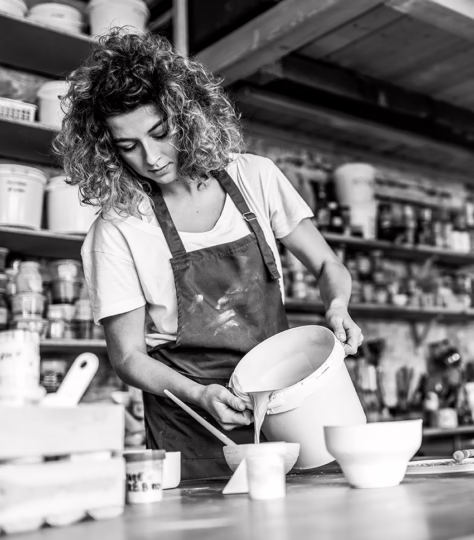 Small business owner wearing a workshop apron pours paint into a bowl with a brush.
