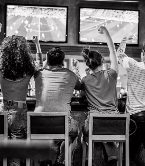 A view of four people from behind, sitting on stools watching sports on TV with their arms cheering in the air.