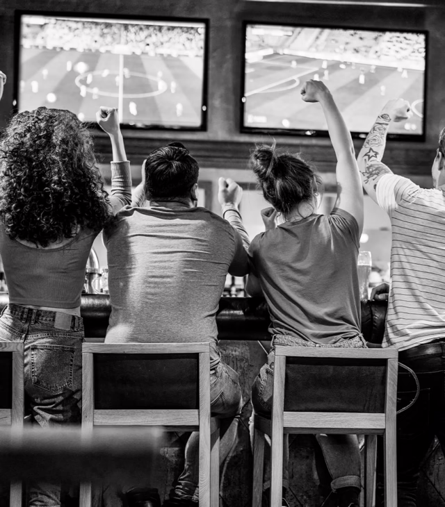 A view of four people from behind, sitting on stools watching sports on TV with their arms cheering in the air.