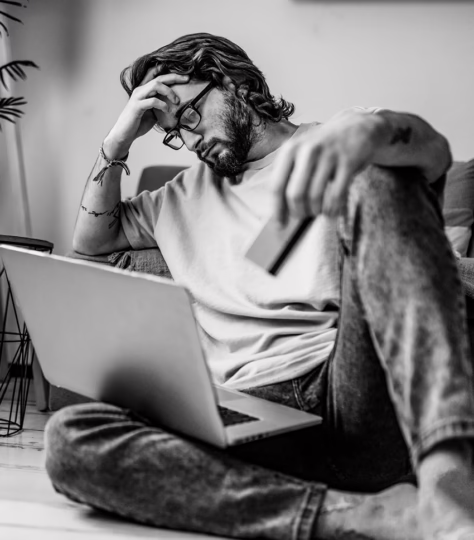 Man with hand over face, on the floor with a laptop in his lap looking upset