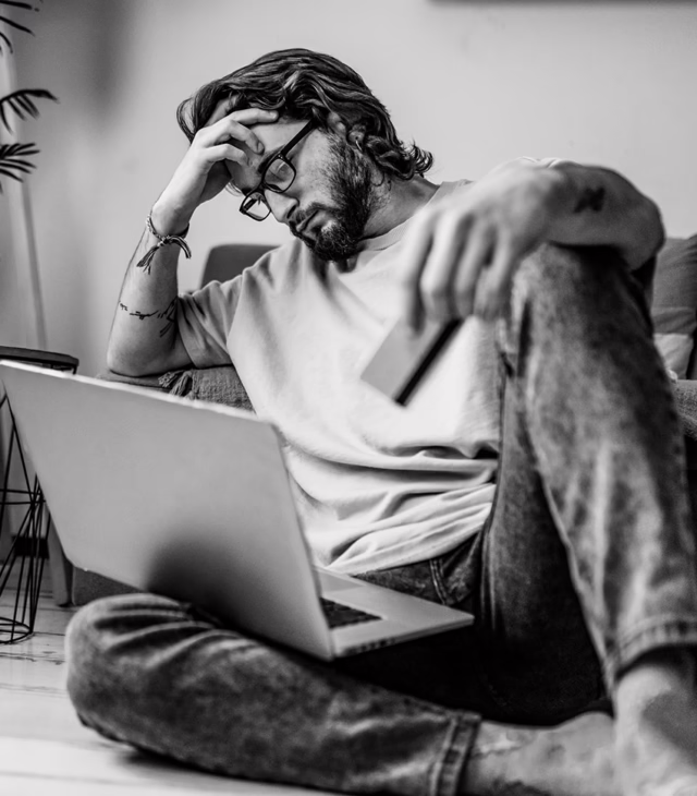 Man with hand over face, on the floor with a laptop in his lap looking upset
