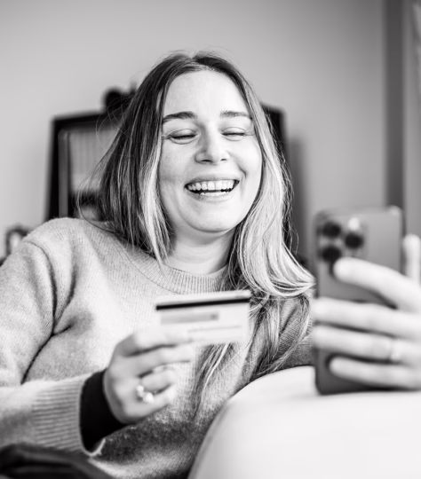 A black-and-white indoor photograph shows a person reclining on a couch while holding a smartphone in one hand and a credit card in the other.